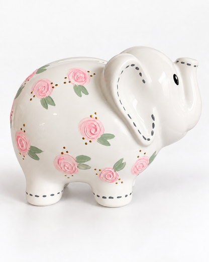 A ceramic elephant-shaped piggy bank with a glossy white finish, decorated with pink roses, green leaves, and dotted black stitch details along its ears, legs, and trunk, set against a plain white background.