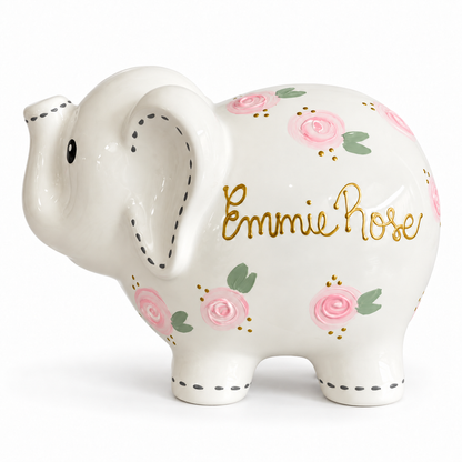 A white ceramic elephant pig bank decorated with pink roses, green leaves, gold cursive text reading 'Emmie Rose', and black dashed line details on the ears and legs, shown on a white background.
