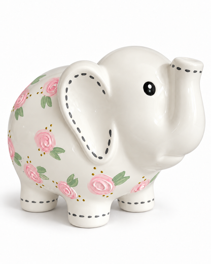 A ceramic elephant-shaped piggy bank in glossy white, decorated with pink roses, green leaves, and black stitched-dot details along the ears, trunk, and legs, shown on a plain white background.