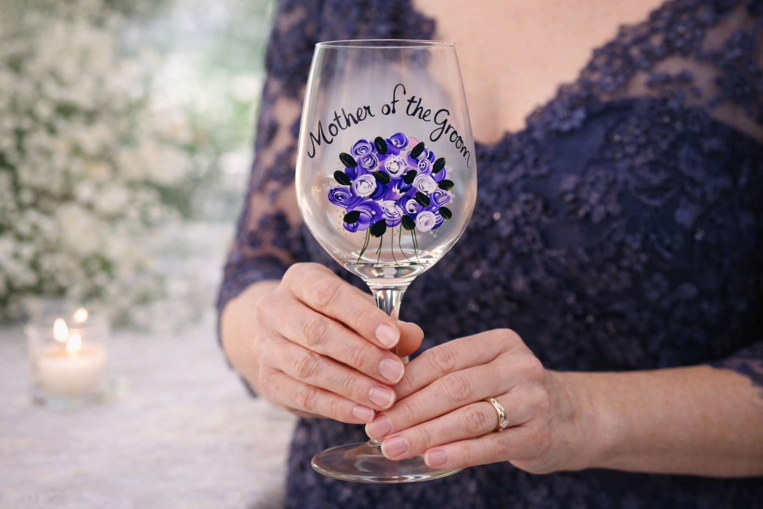 Person holding a wine glass with 'Mother of the Groom' and floral design, blurred floral background