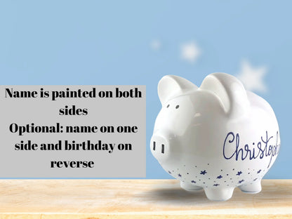 White piggy bank with 'Christopher' painted on it, placed on a wooden surface with a light blue background.